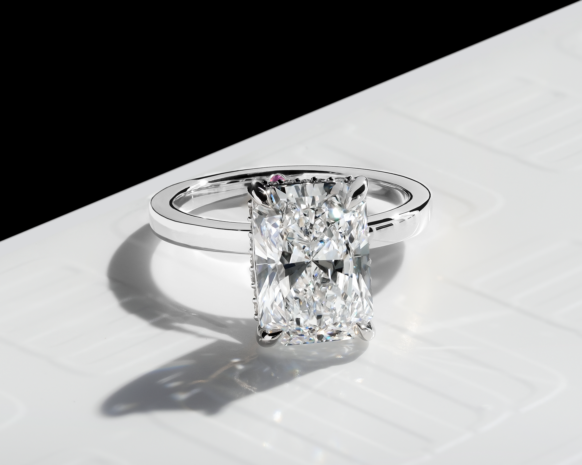 Close-up view of a radiant-cut diamond engagement ring displayed on a polished white surface with the MJ Jones monogram subtly embossed. The ring showcases a stunning radiant-cut diamond with sharp corners, reflecting brilliance and clarity. A hidden pink diamond accent is visible beneath the center stone, adding a touch of uniqueness. The composition highlights the diamond’s clean facets and sparkle, complemented by a sleek & polished band.